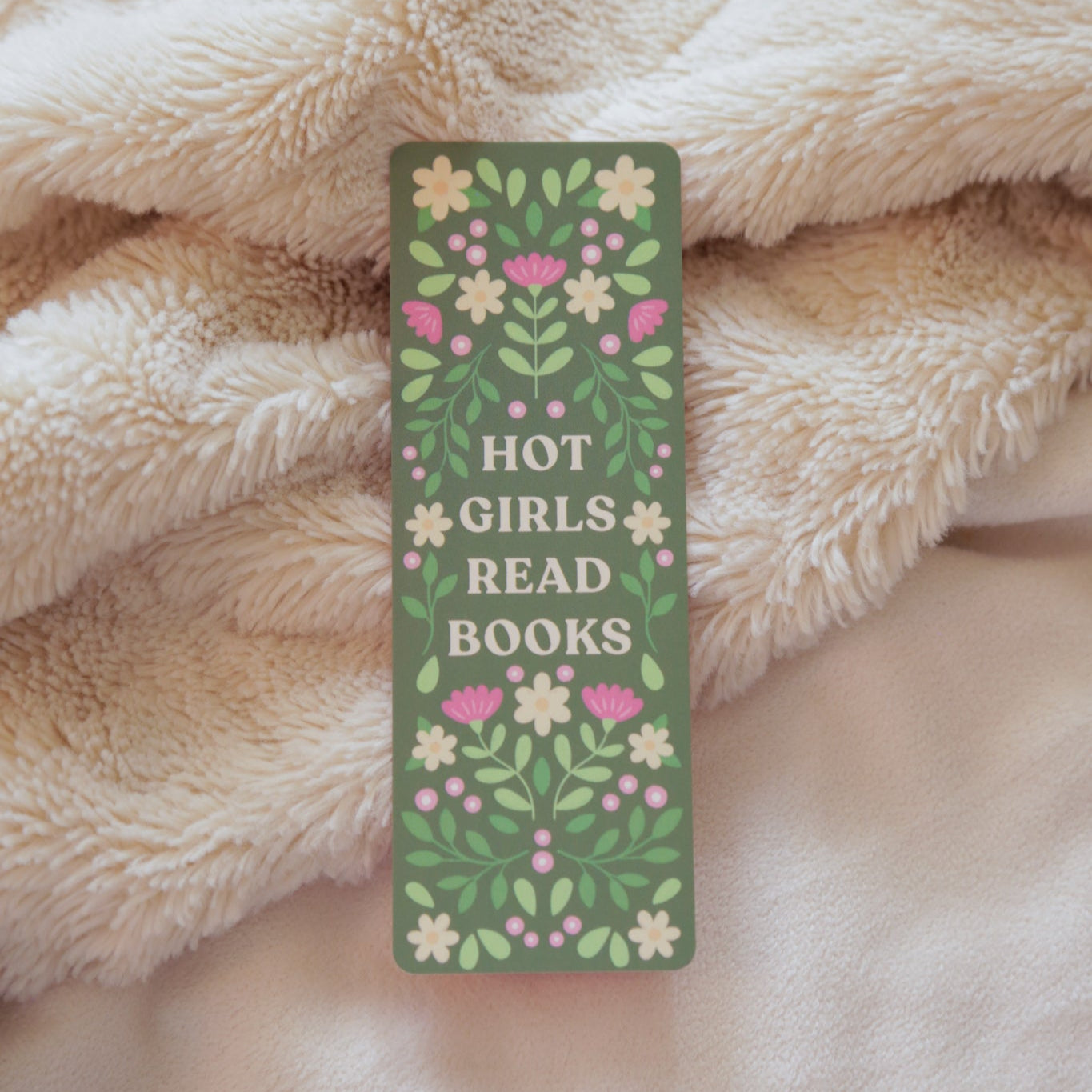 A photograph of a green floral bookmark with the quote "hot girls read books", resting against some bedding.