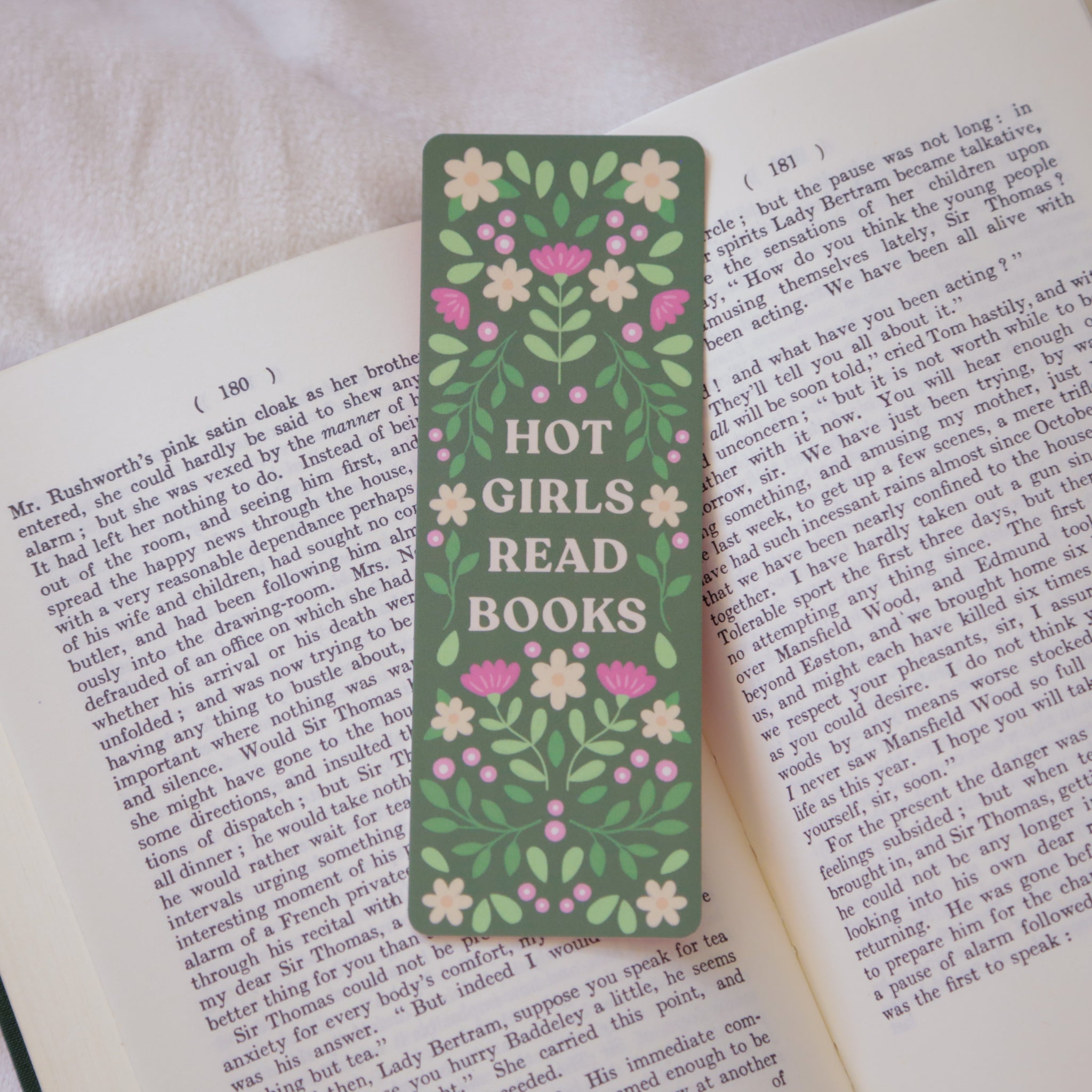 A photograph of a green bookmark against an open book, the bookmark reads "Hot Girls Read Books".