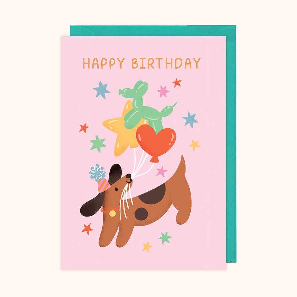 A colorful birthday card featuring an illustration of a dog with balloons, accompanied by text 'Happy Birthday'. The card is accompanied by a teal envelope.