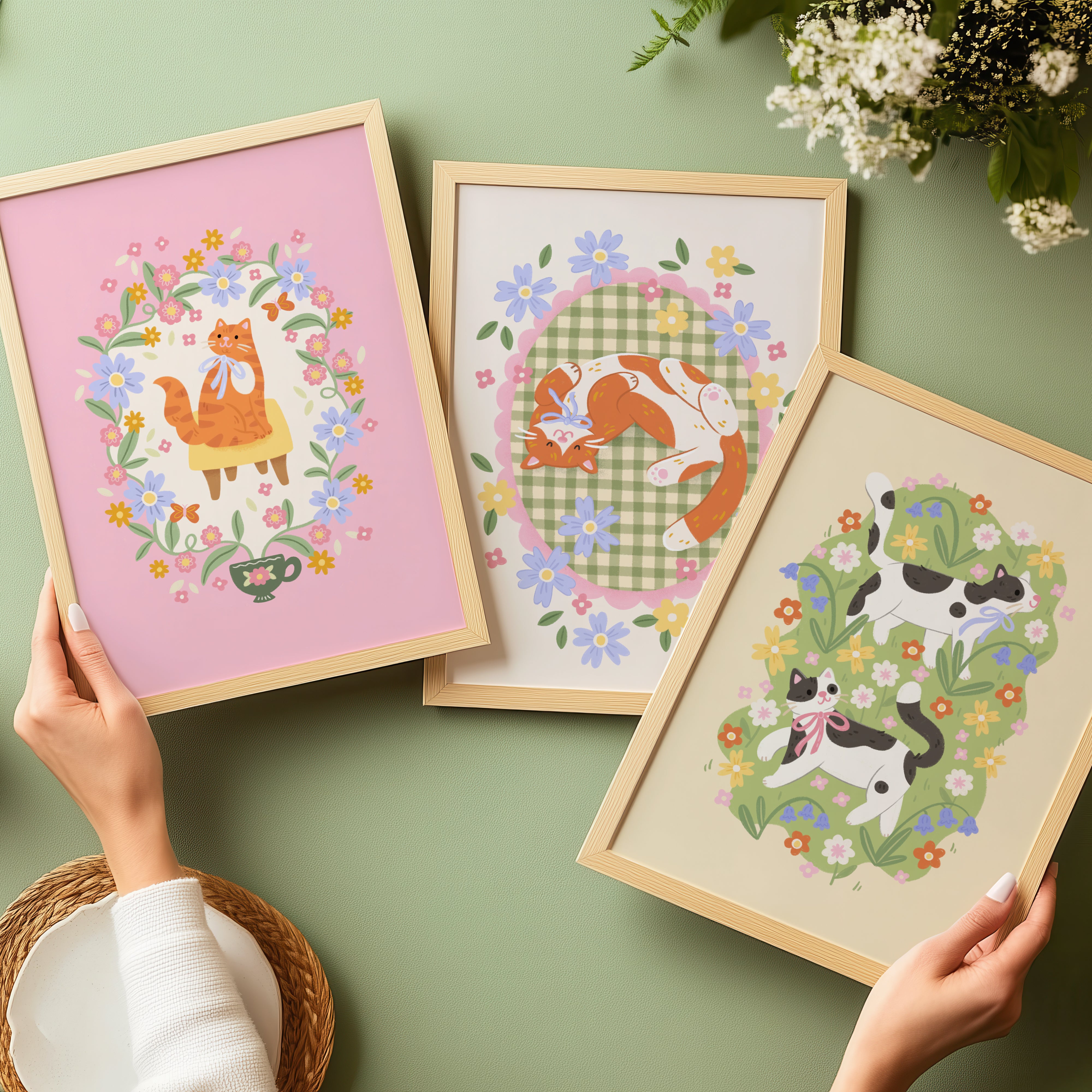 Three framed illustrations of cats with floral wreaths on a green surface.