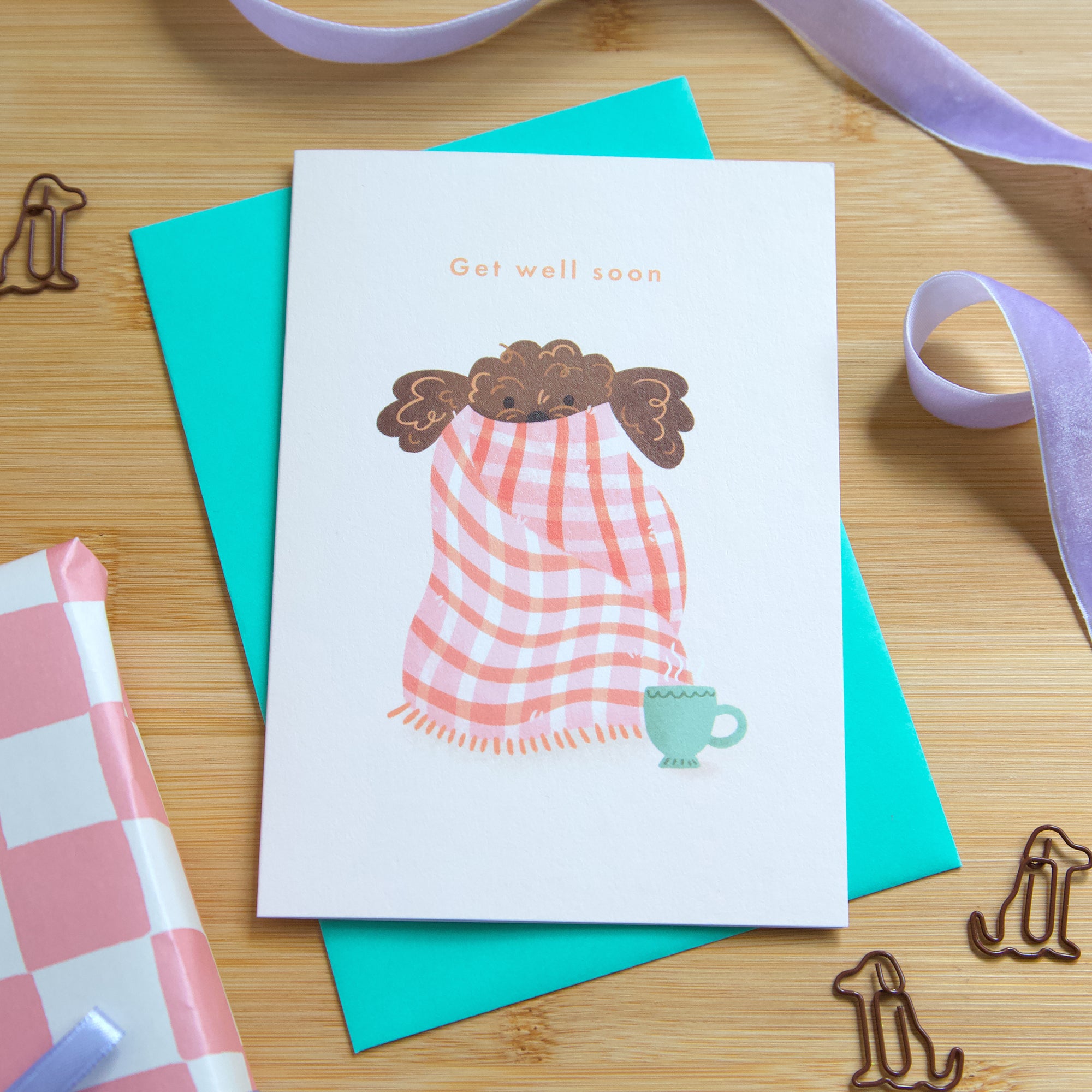 A photograph of a get well soon greetings card featuring a puppy wrapped in a blanket with a cup of tea. 